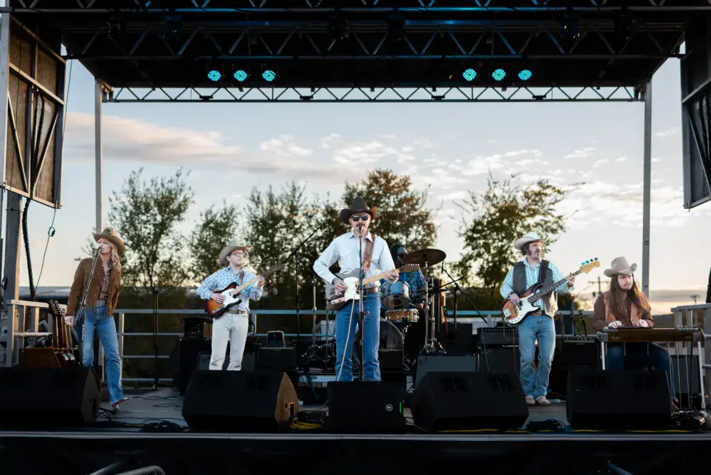 A five-piece country band performs on an outdoor stage with trees and sky in the background.