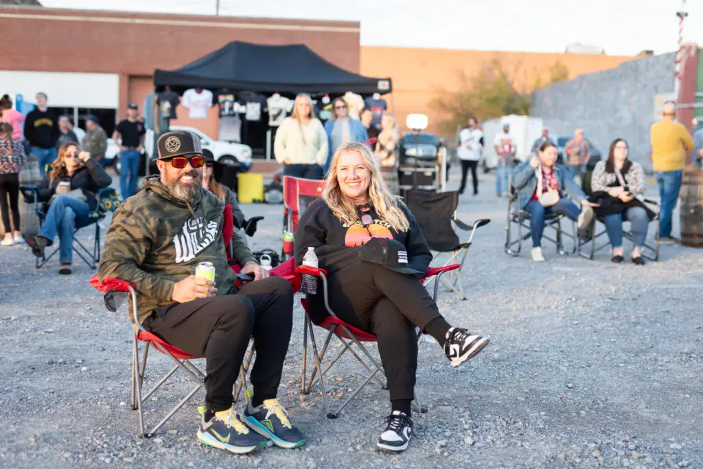 Two smiling attendees sit in red camp chairs in the foreground at an outdoor music festival venue.