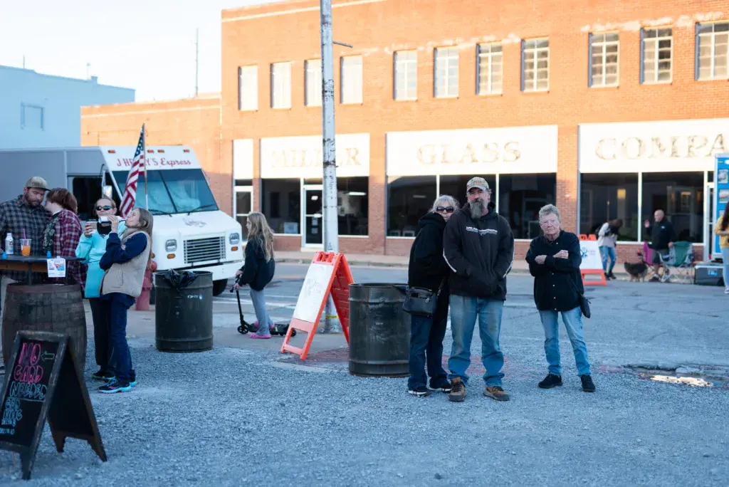 Festival attendees gather in a downtown street in front of brick buildings near a food truck and vendor area.