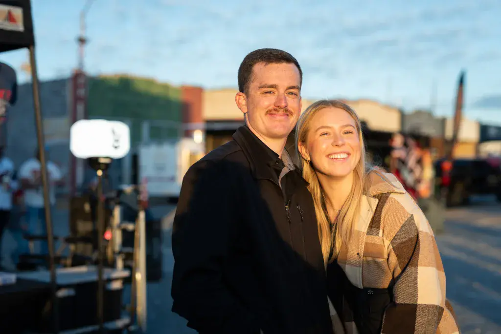 Two people smiling at the festival with venue structures and crowd visible in the background.