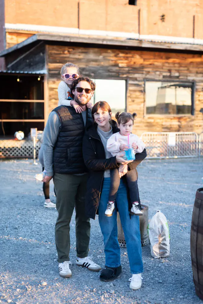 A family of four smiles at the camera in front of wooden venue buildings at the festival.