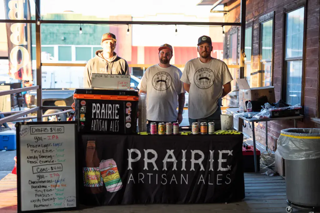 Three staff members stand behind a Prairie Artisan Ales booth with beer samples and menu board displayed.