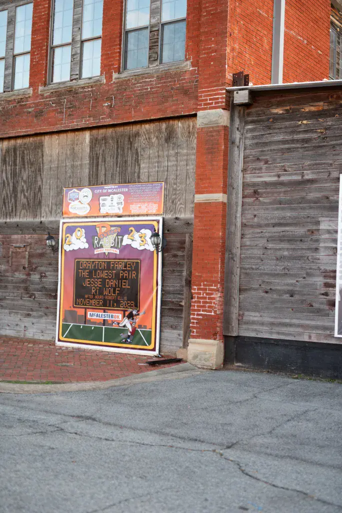 Festival signage posted on a weathered wooden wall of a brick building announcing upcoming performances.