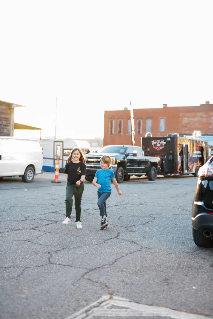 Two children running through a festival parking lot with vintage buildings and parked vehicles in the background.