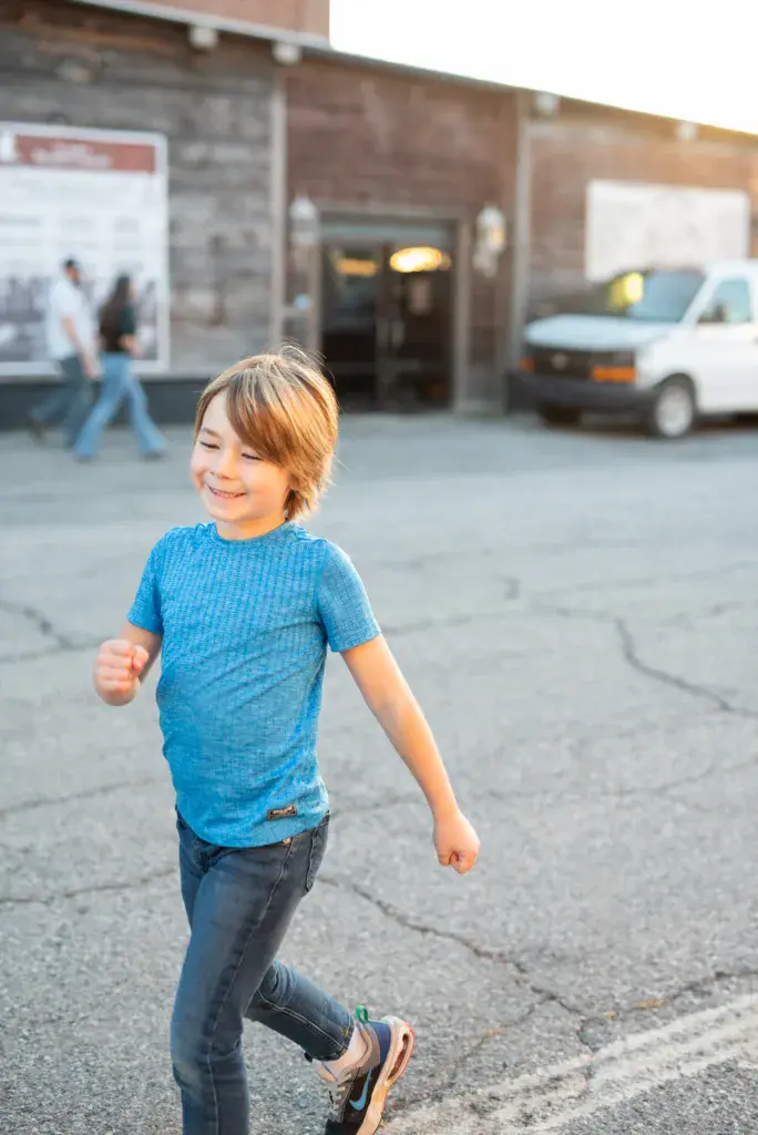 Young child in blue shirt running and smiling in a parking lot during daytime.
