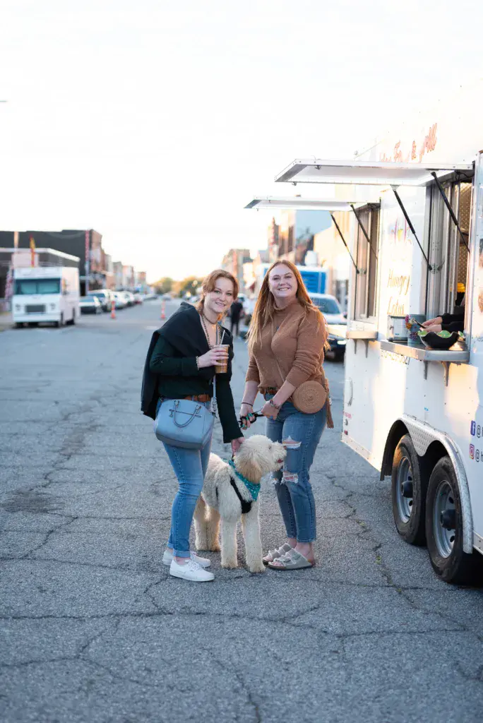 Two people with a dog stand smiling in front of a food truck at the festival venue.