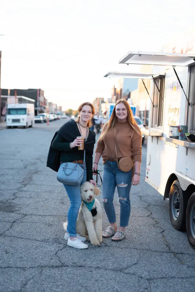 Two people and a white dog stand on a street next to a food truck at dusk.