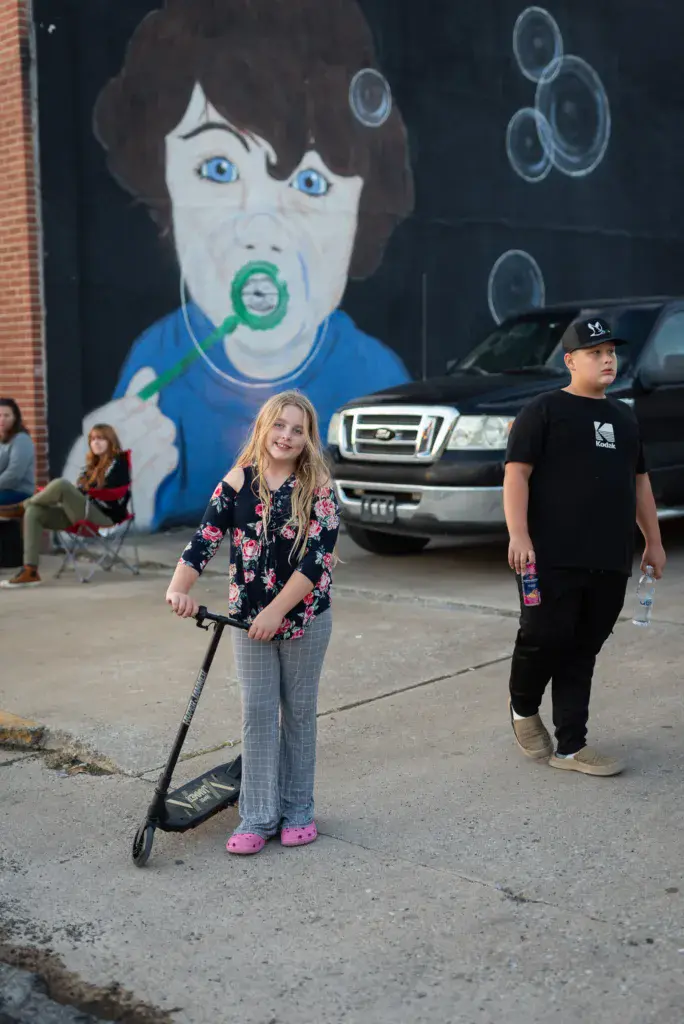 Festival attendees gather in a parking lot near a large mural of a person with blue eyes and green circular mouth design.