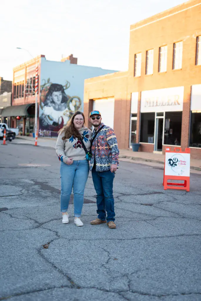 Two people pose together on a city street in front of historic brick buildings with colorful murals and storefront signage.