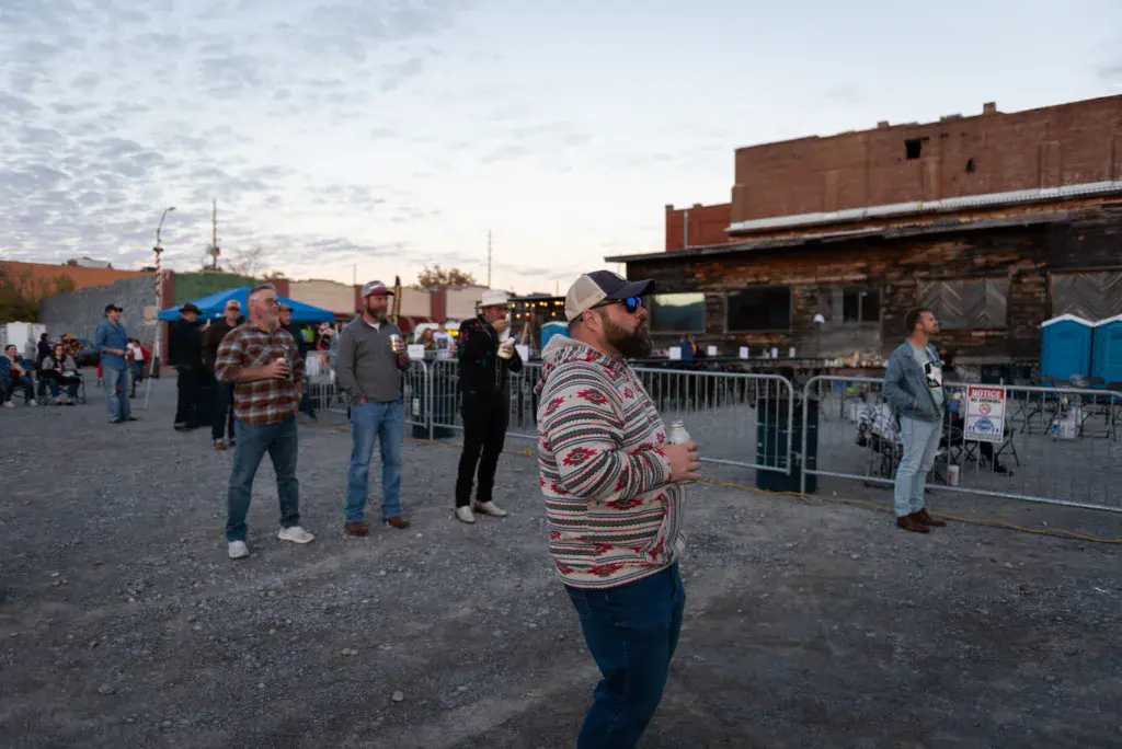 Attendees gather in an outdoor festival venue near brick buildings at dusk.