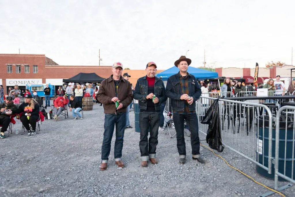 Three men in casual attire walk together through a festival grounds with crowds and vendor tents in the background.