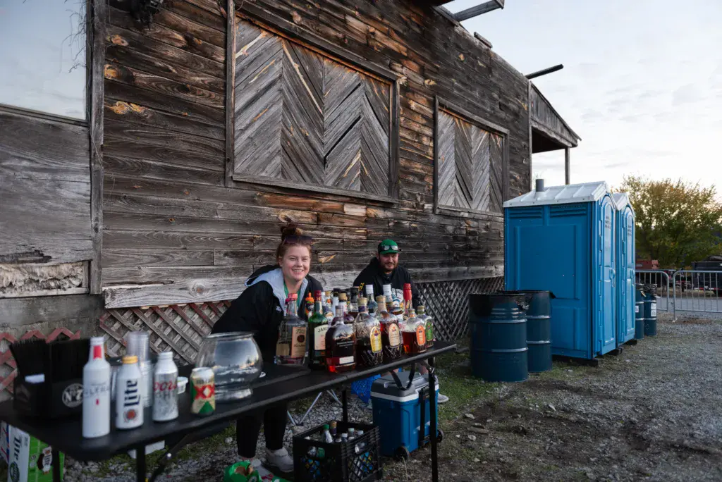 Two people staffing a beverage stand with bottles and cans displayed on a table in front of a rustic wooden structure.