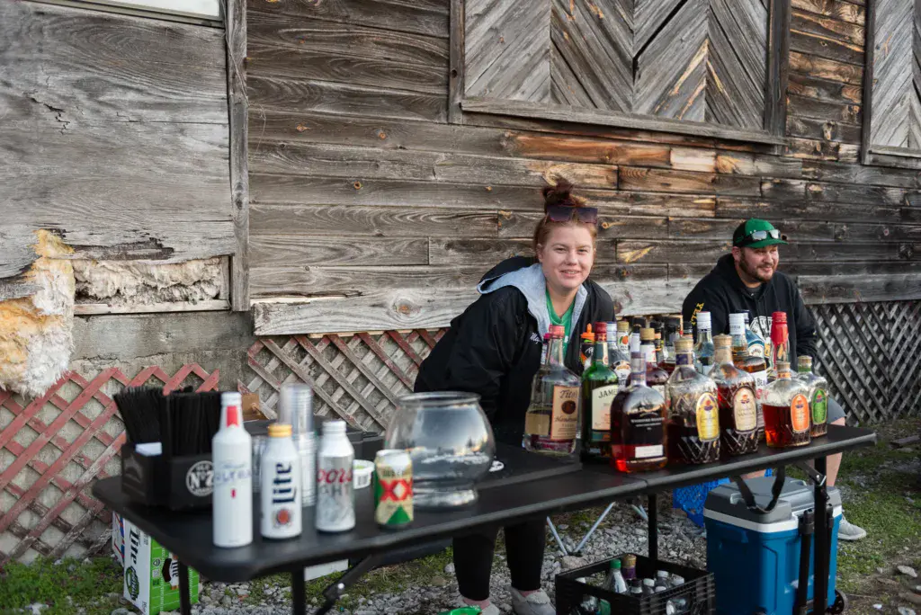 Two people operate a beverage vendor stand with bottles and cups at an outdoor festival.