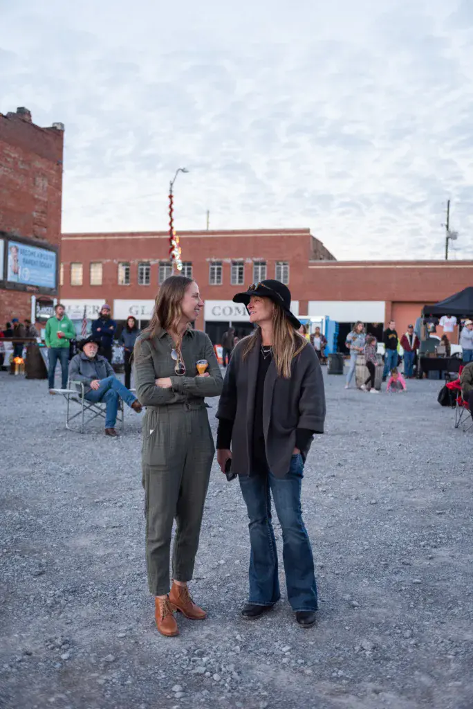 Two people converse in a festival ground with brick buildings and crowds visible in the background.