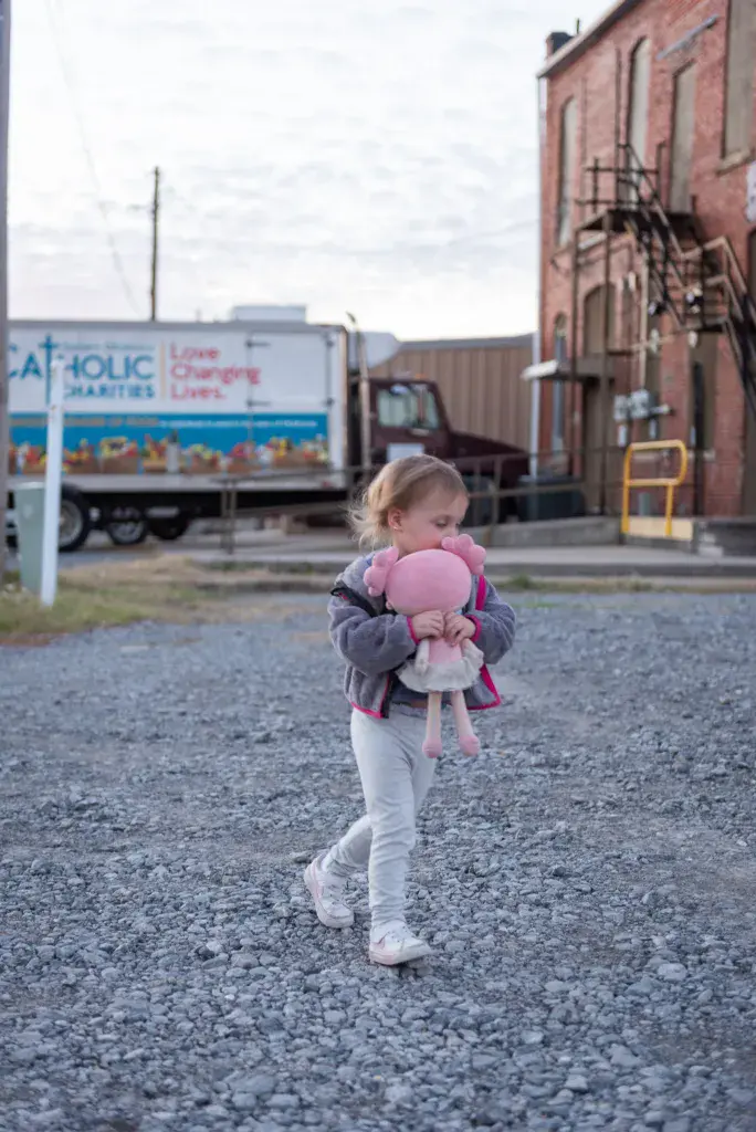 A young child holds a pink stuffed animal while walking through a festival venue near industrial buildings and a food truck.