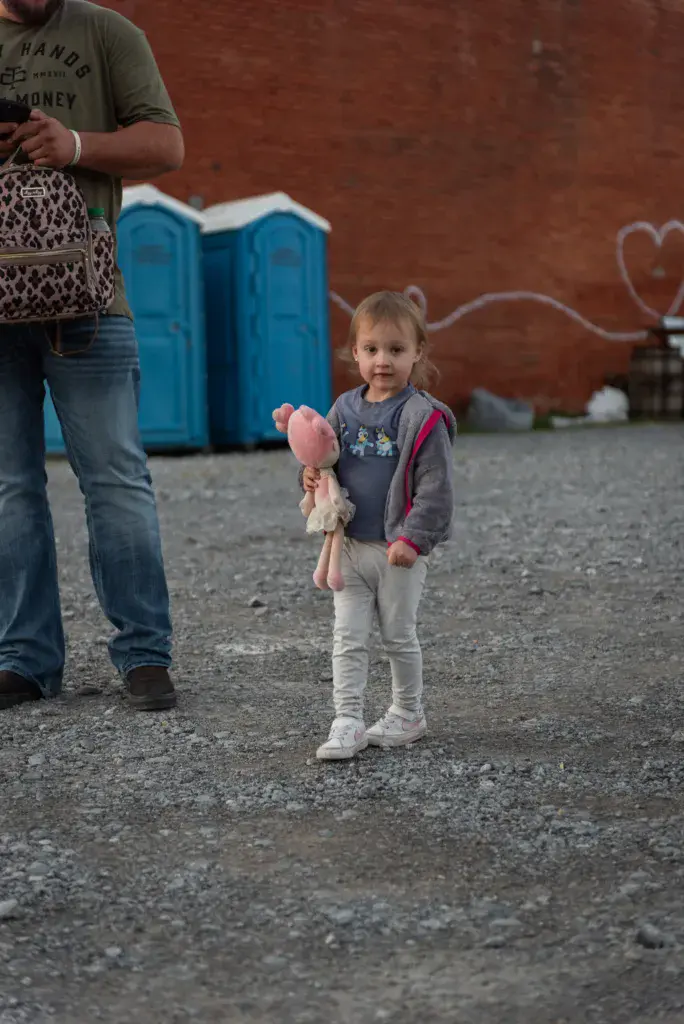 A young child holding pink cotton candy stands near an adult at a festival venue with portable toilets visible in the background.
