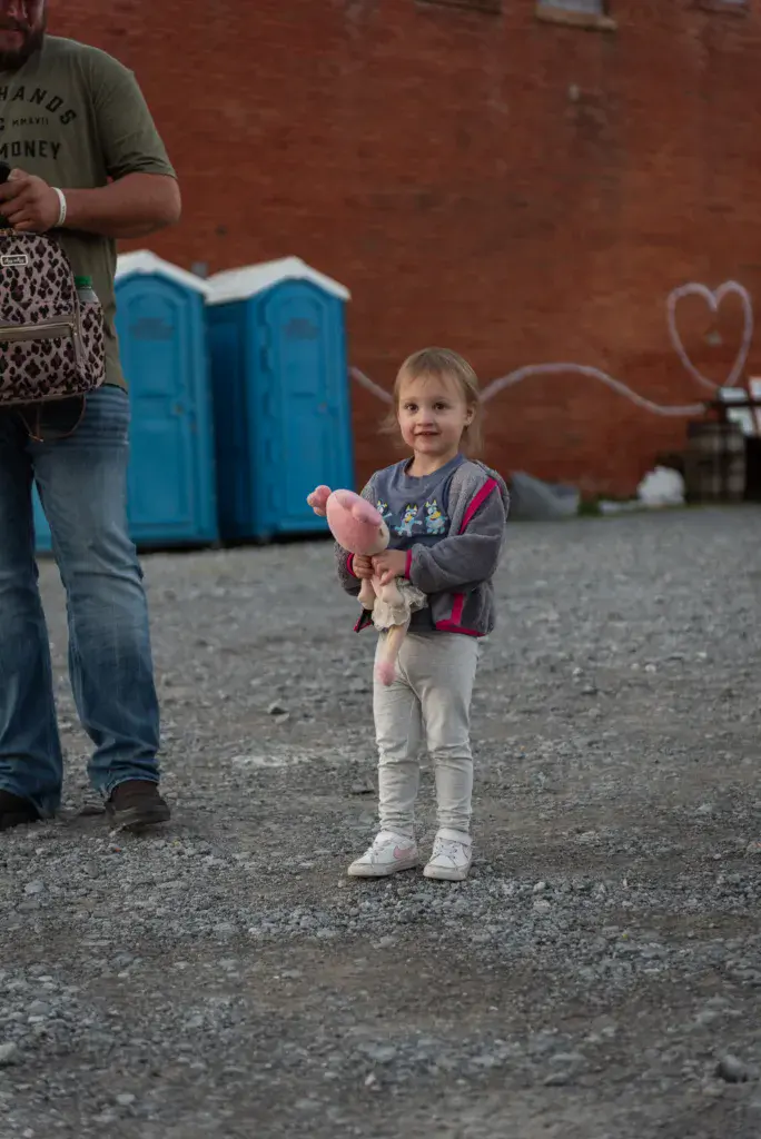 A young child holds cotton candy while standing in a festival venue near portable restrooms and a brick wall.