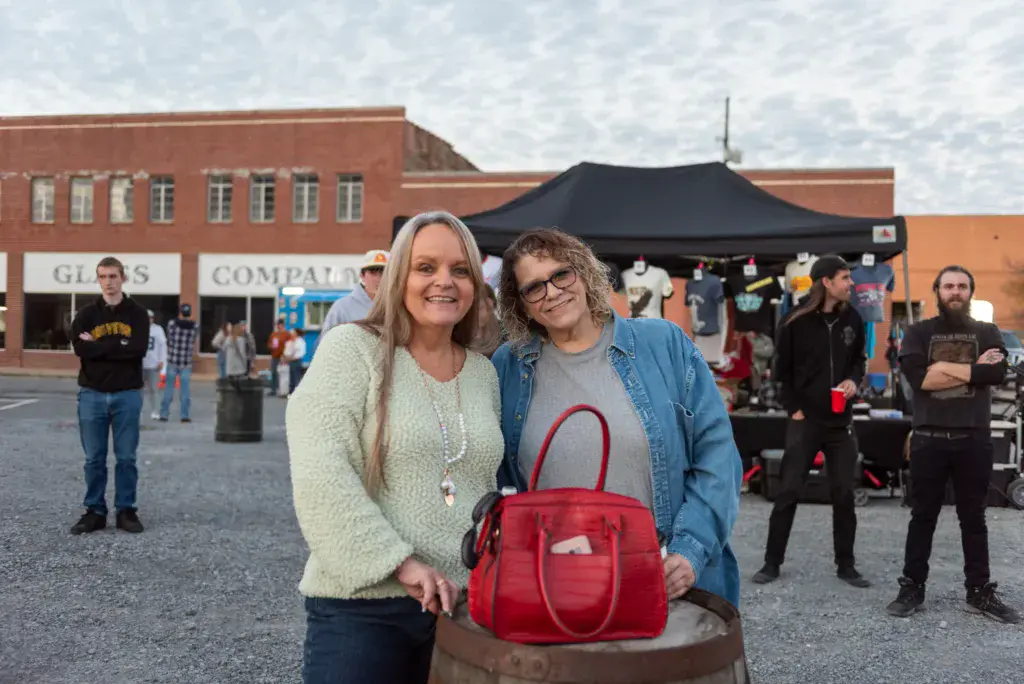 Two women smile at the camera at the Dancing Rabbit Music Festival in a town square with brick buildings and a black tent in the background.