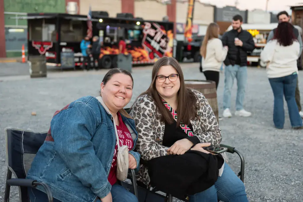 Two smiling attendees sit together at the Dancing Rabbit Music Festival with food vendors and other guests visible behind them.