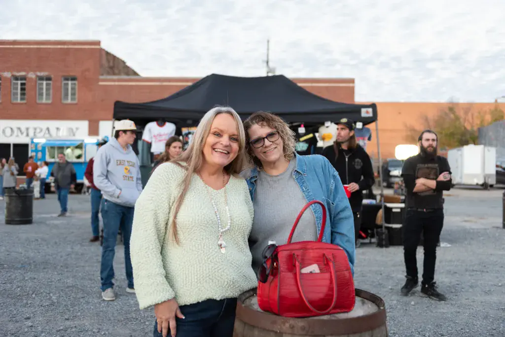 Two smiling women pose together at an outdoor festival venue with brick buildings and a black tent in the background.