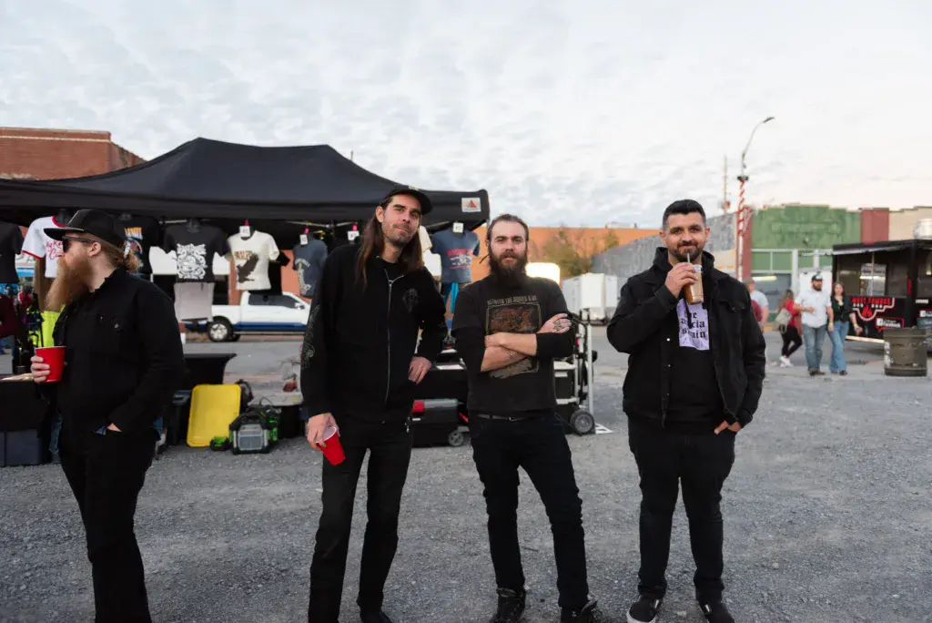 Four men in dark clothing stand together at an outdoor festival venue with tents and crowds visible behind them.