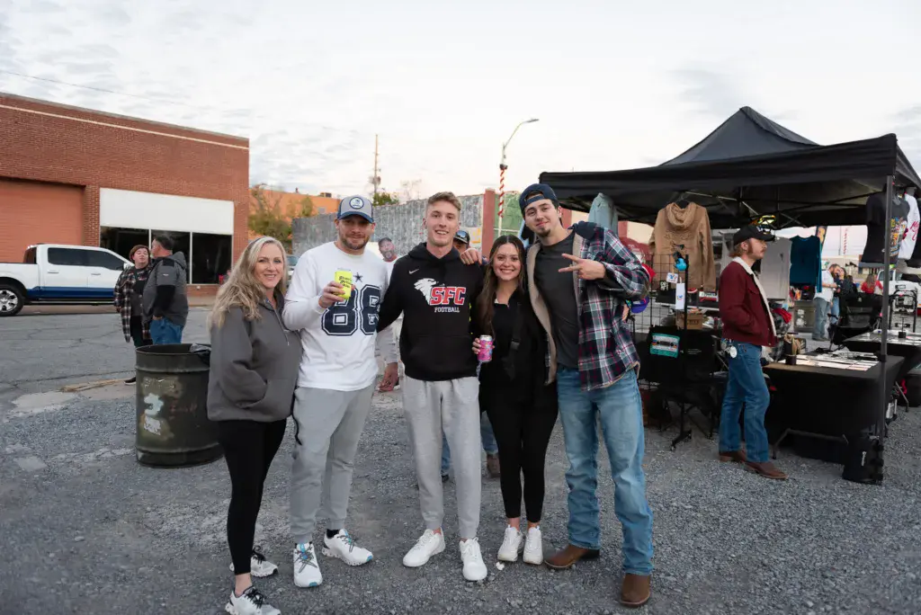 A group of five people pose together at the Dancing Rabbit Music Festival, with vendor tents and festival grounds visible behind them.