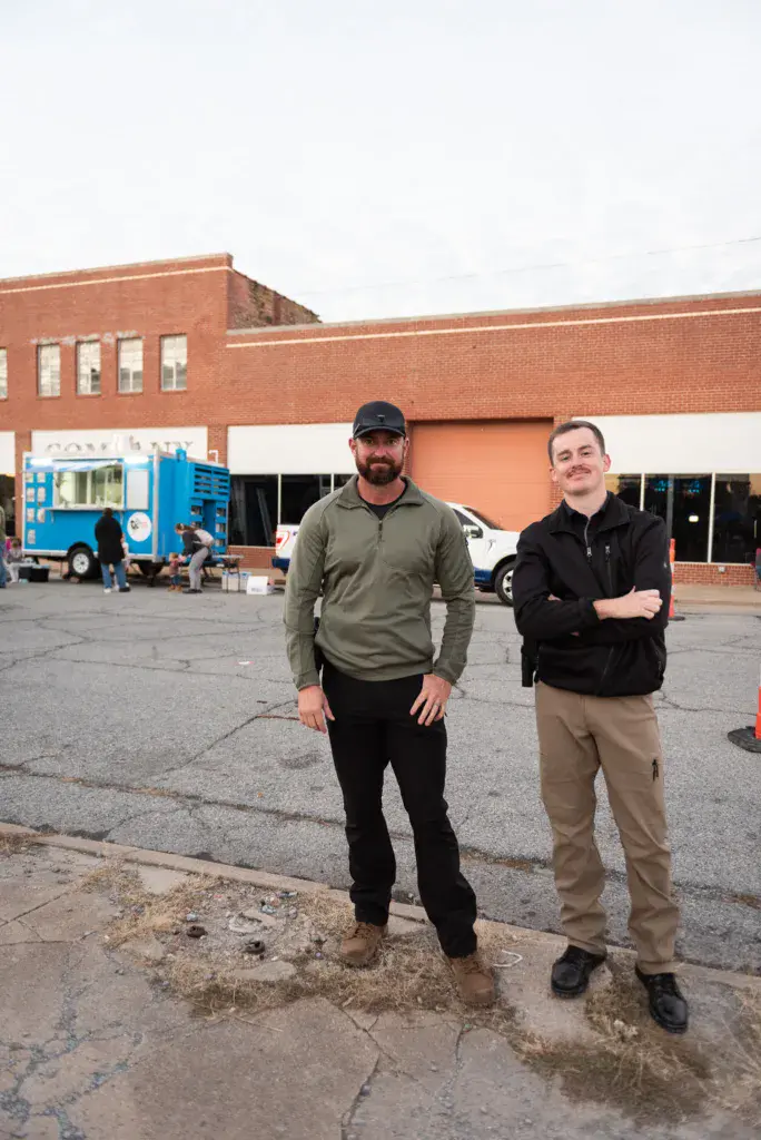 Two people stand in a parking lot in front of a brick building with a blue food truck visible behind them.