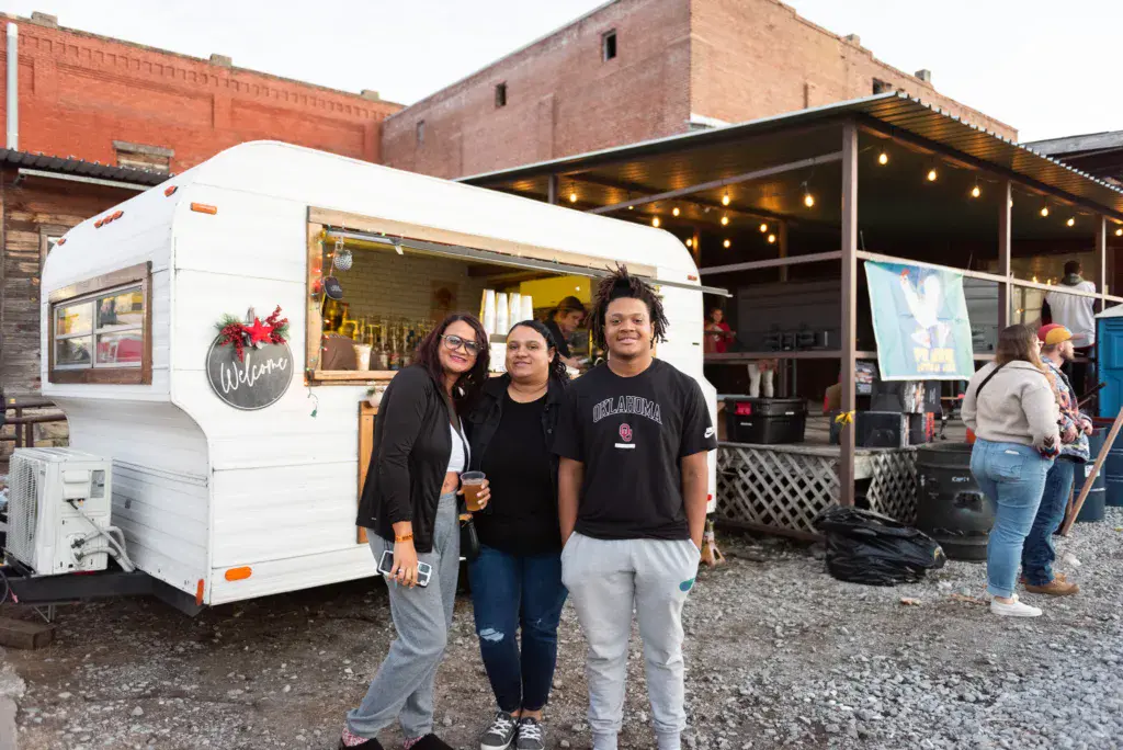 Three people pose together in front of a white food vendor trailer at an outdoor festival venue.