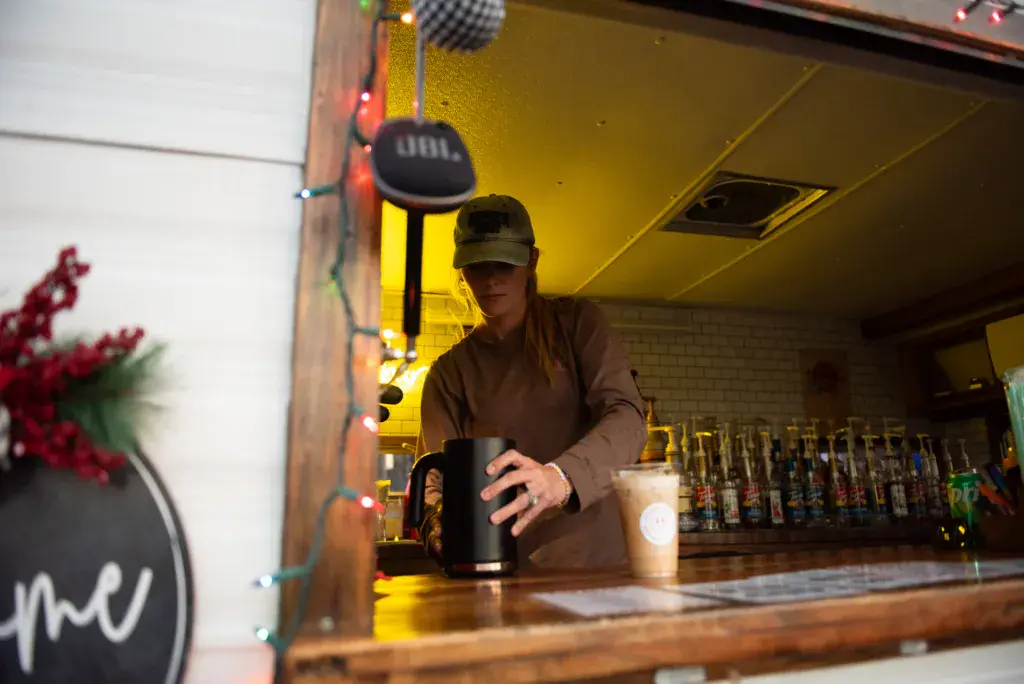 A person in a brown jacket and cap stands behind a festival bar counter, holding a black mug near beverage dispensers.