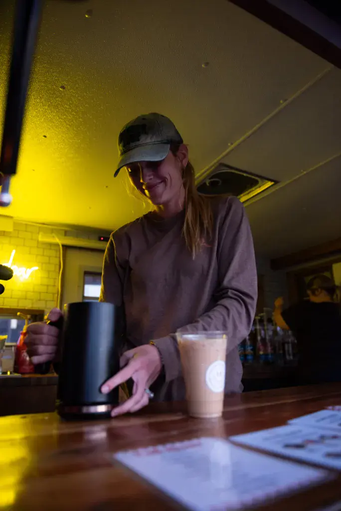 A person in a visor smiles while serving drinks at an indoor venue with yellow lighting.