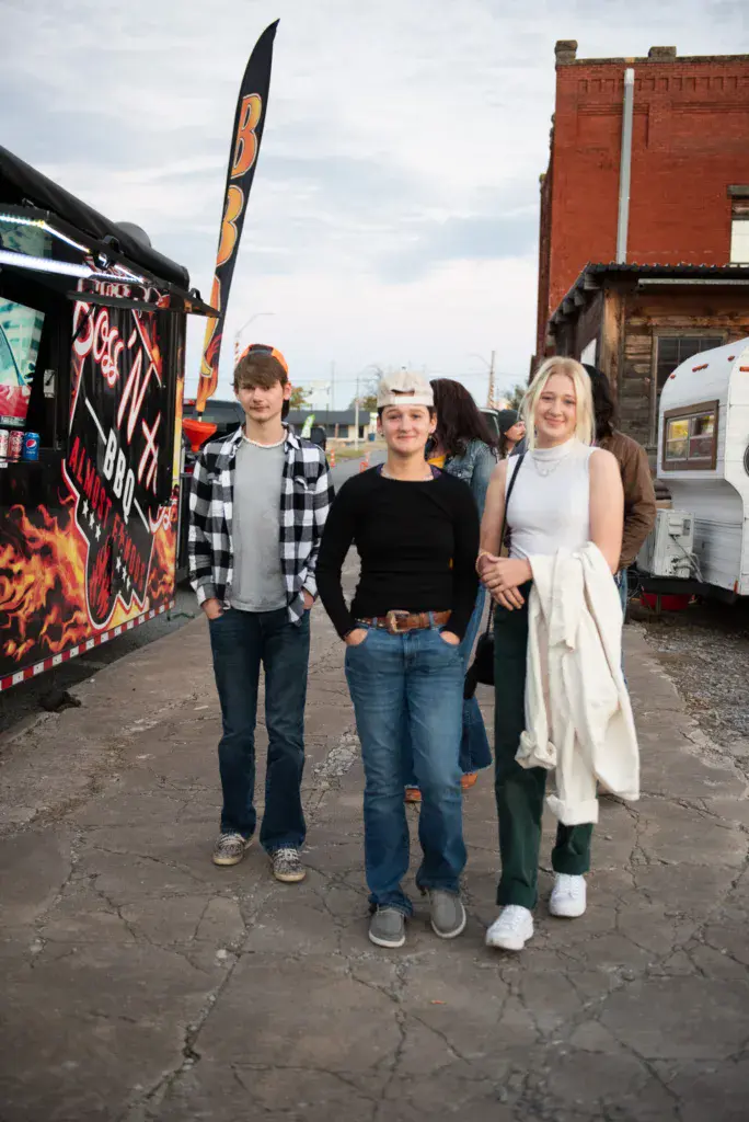 Three festival attendees pose together in front of vendor booths and brick buildings at the outdoor event.