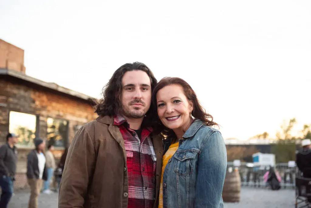 Two people pose together for a portrait at an outdoor festival venue with a stone building and other attendees in the background.