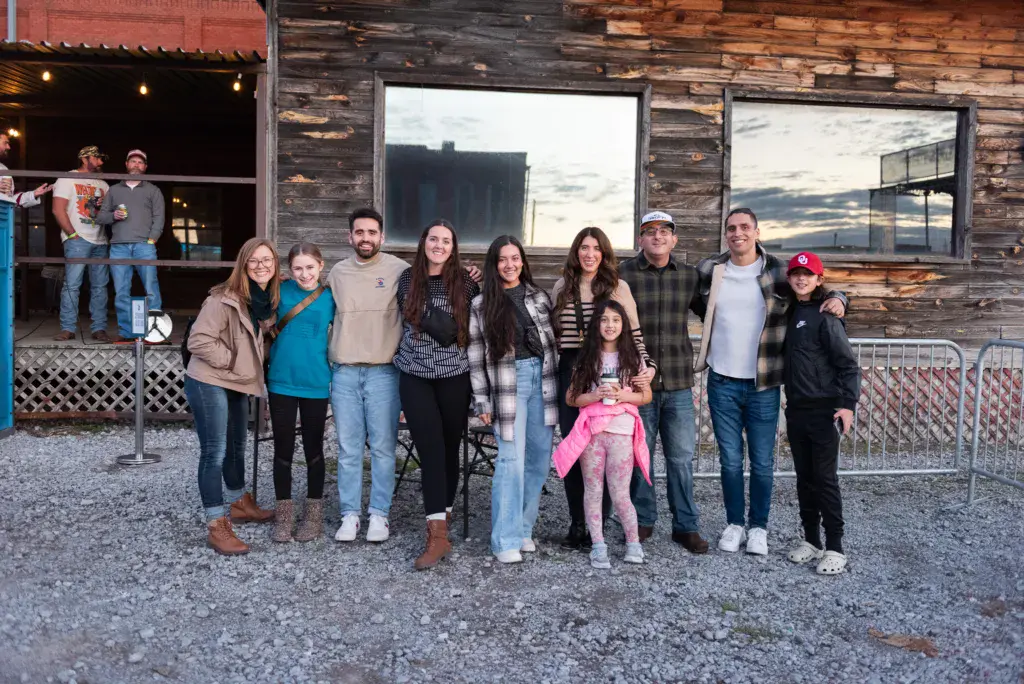 A large group of people poses together in front of a rustic wooden venue building at dusk.