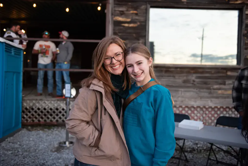 Two young people smile at the camera inside a rustic venue with festival attendees visible in the background.