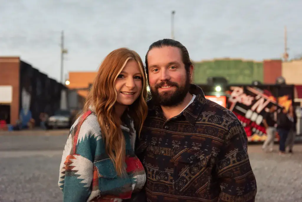 Two people pose together at an outdoor festival venue with colorful buildings and crowds visible in the background.