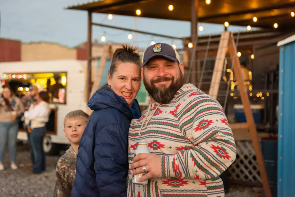 A family of three smiles at the camera in front of a decorated festival venue with string lights at dusk.