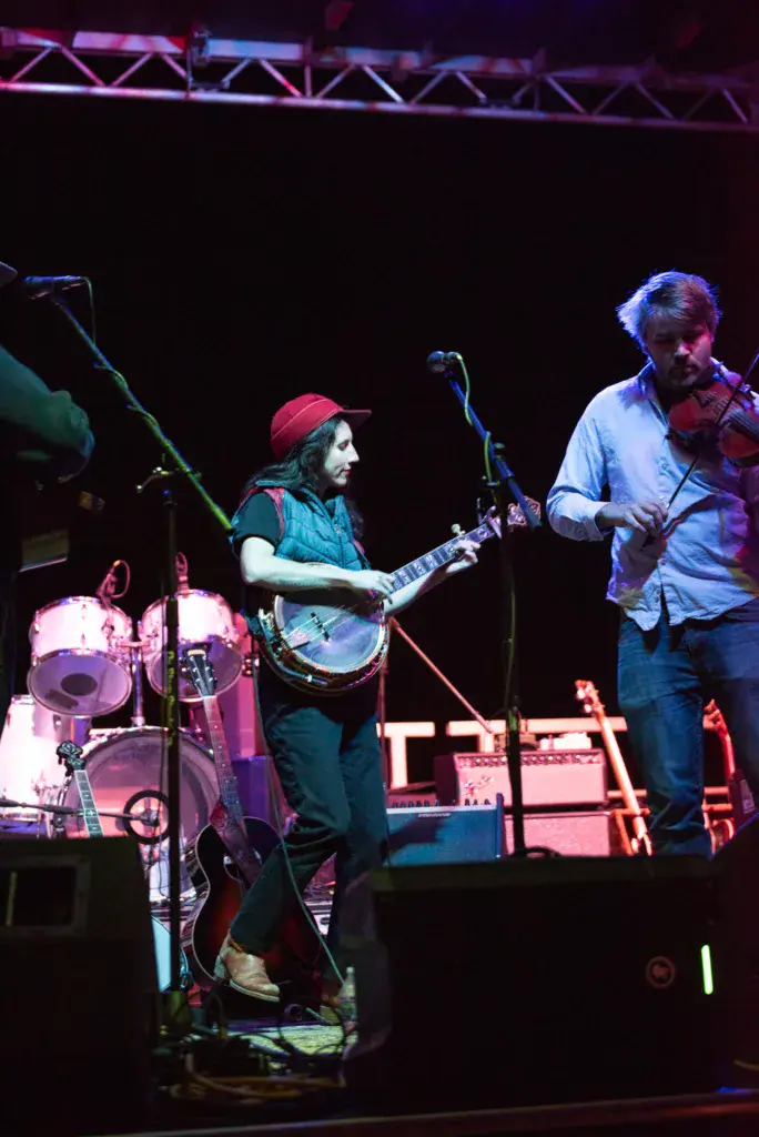 Two musicians perform on stage under purple lighting, one playing banjo and one playing violin.