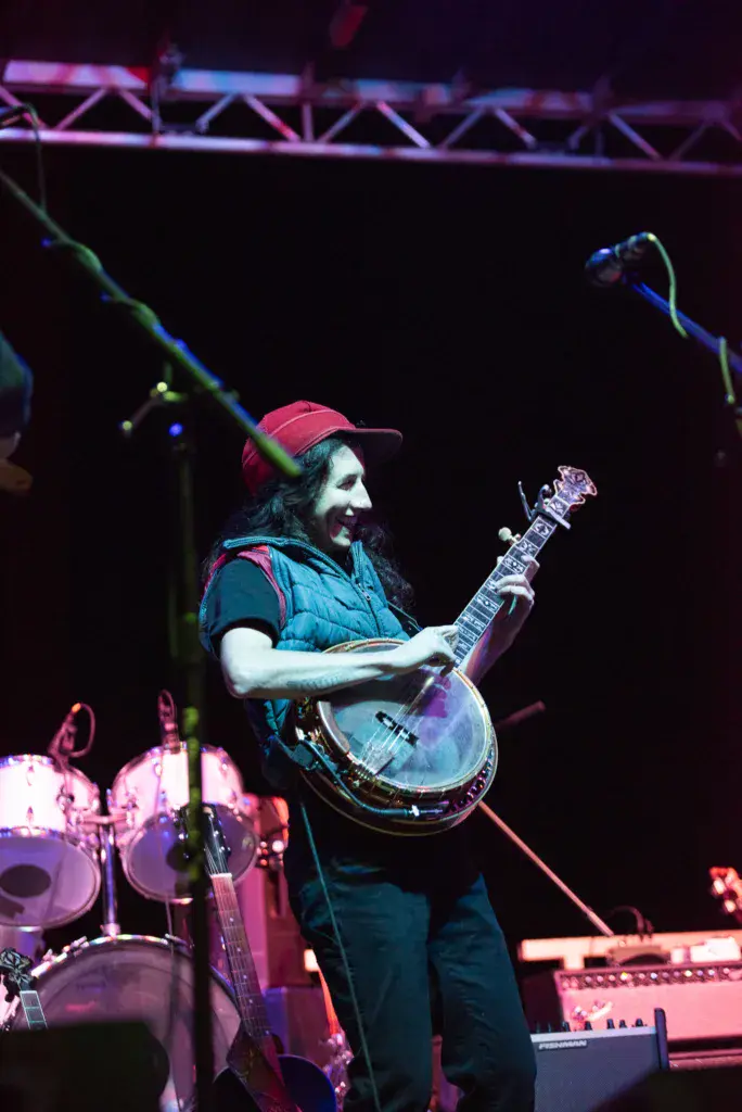 A musician wearing a red cap and blue vest plays a banjo on stage under colorful lighting at an outdoor festival.