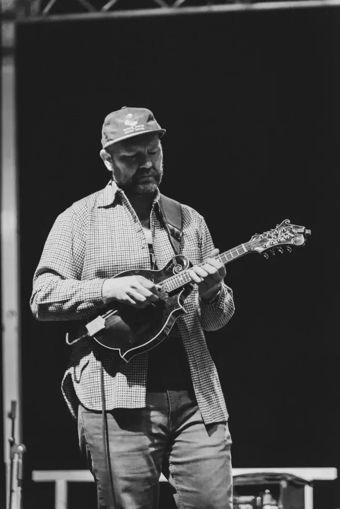 A musician performs on stage wearing a checkered shirt and cap while playing a mandolin against a dark backdrop.