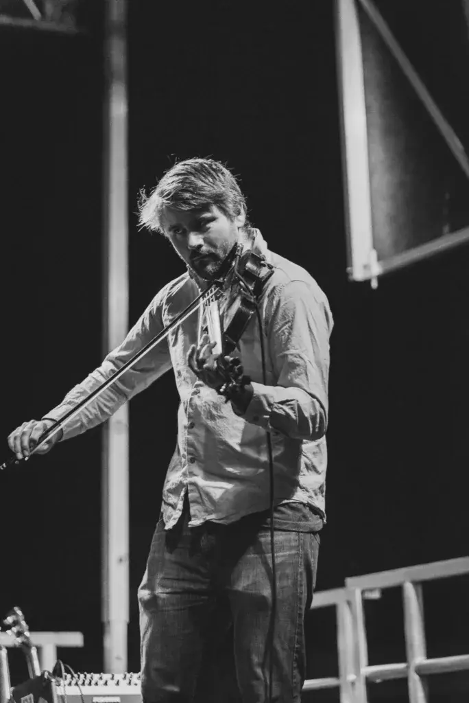 Musician with headphones around neck playing electric violin on stage with stage lighting in background.