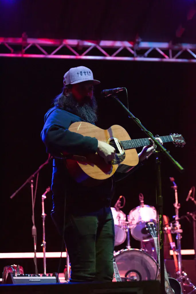 A bearded performer wearing a cap plays an acoustic guitar at the Dancing Rabbit Music Festival stage.