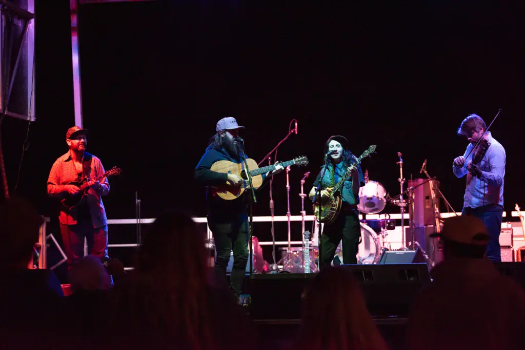 Five musicians perform on stage with guitars and other instruments under purple and red stage lighting.