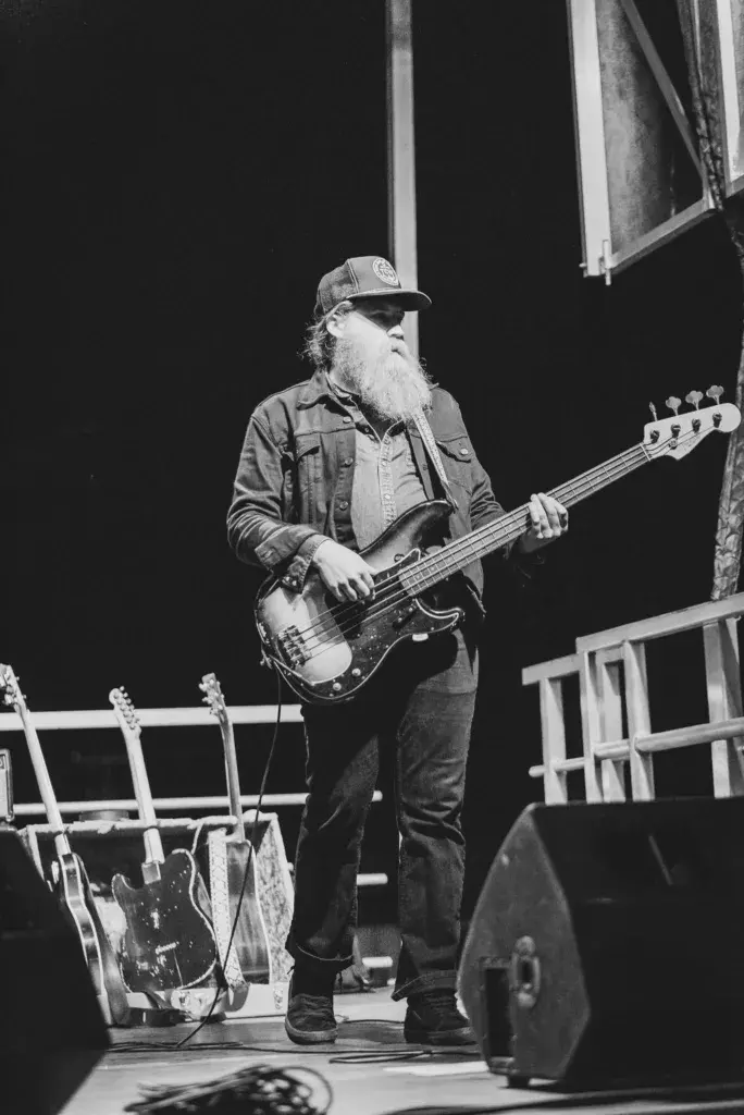 Bassist with long beard and cap performing on stage under stage lights at night.