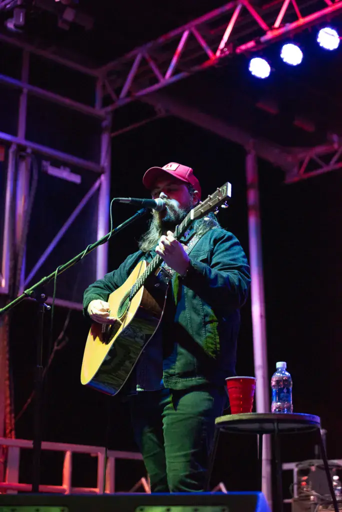 A bearded performer wearing a red cap plays an acoustic guitar and sings into a microphone on an illuminated stage.