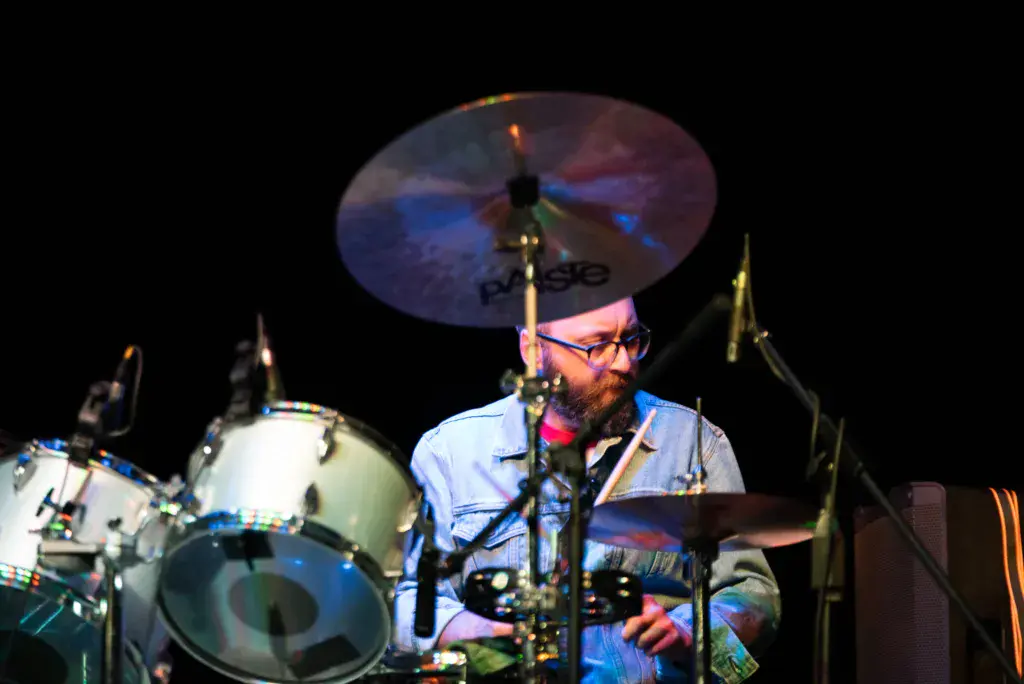 A drummer wearing glasses performs on stage at night, playing a drum kit with colorful stage lighting.
