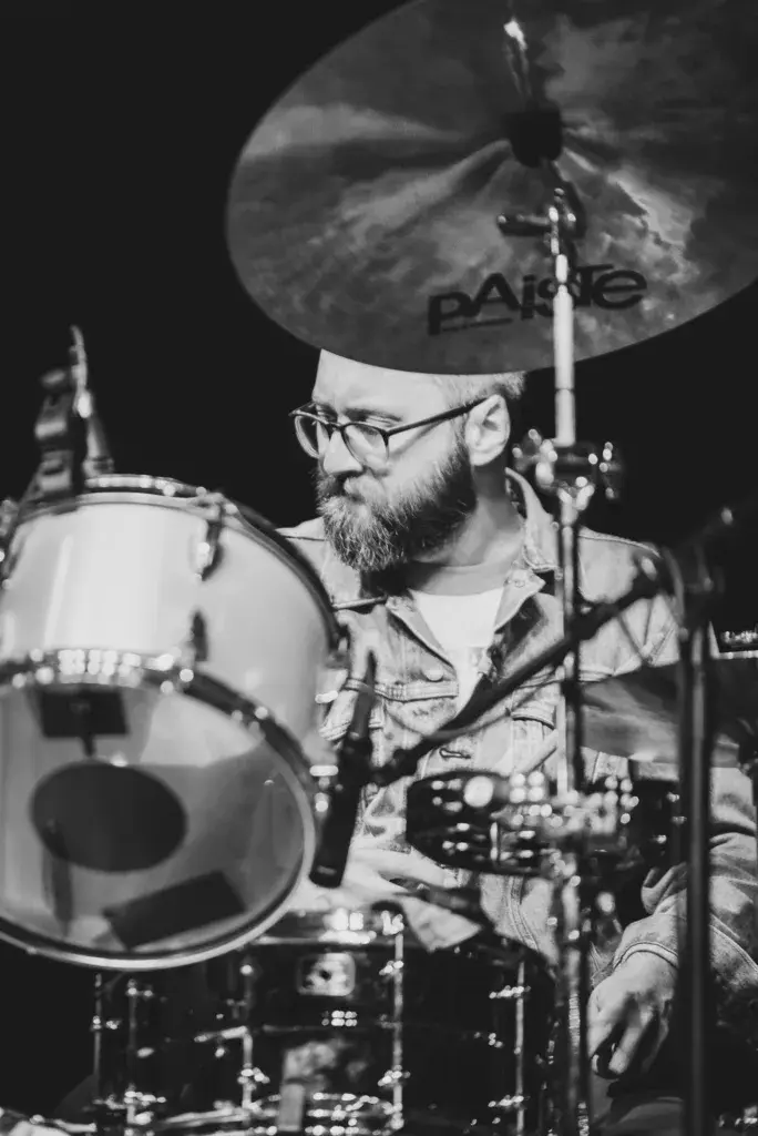 A drummer with glasses and a beard plays behind a Paiste cymbal during a nighttime performance.