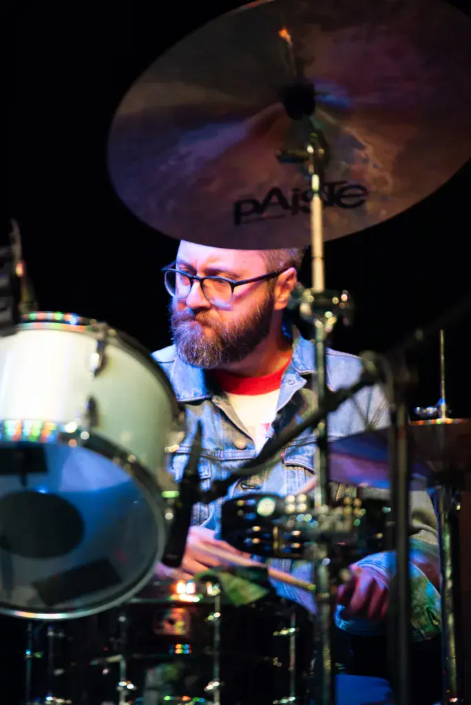 Drummer wearing glasses and red shirt performs on stage with colorful cymbals and drums.