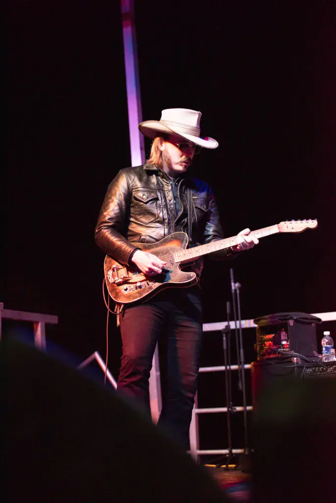 A guitarist in a leather jacket and cowboy hat performs on stage under purple stage lighting.