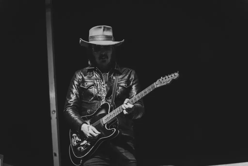 A musician wearing a cowboy hat and leather jacket plays an electric guitar on stage in black and white.