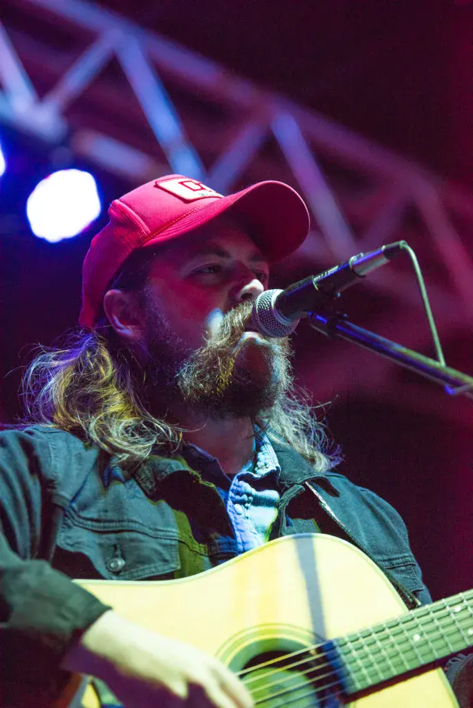 A bearded musician wearing a red cap performs with an acoustic guitar at the festival stage.